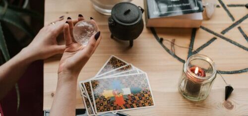 From above of crop unrecognizable female soothsayer with magic ball near tarot cards and candles during divination process in house