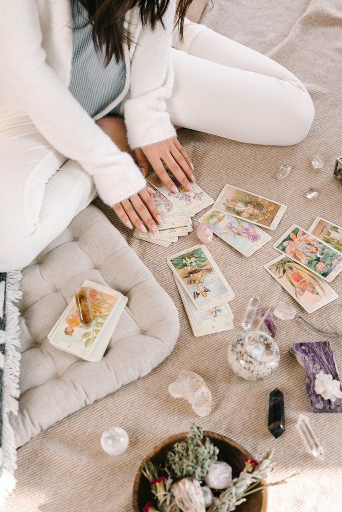 A woman sits cross-legged indoors, handling tarot cards, surrounded by crystals and herbs.
