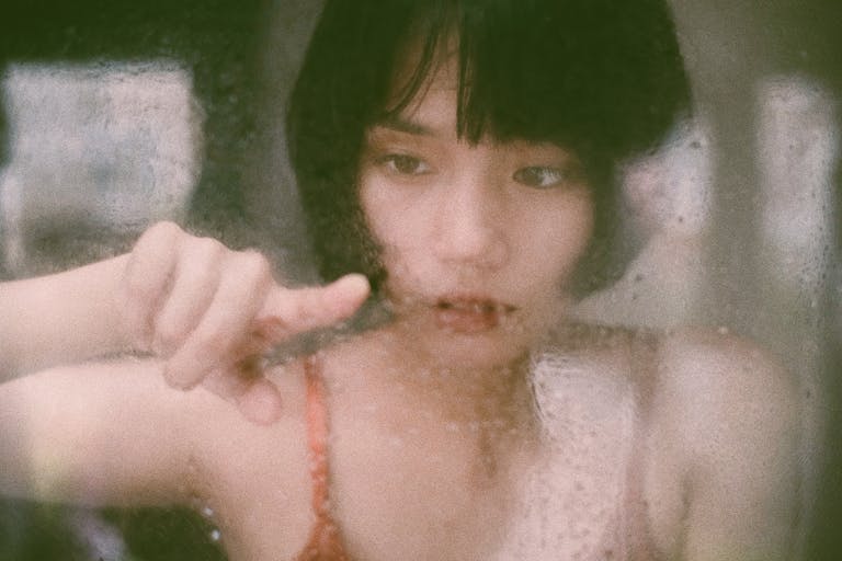 Through window of young Asian woman drawing on wet glass with finger in soft light